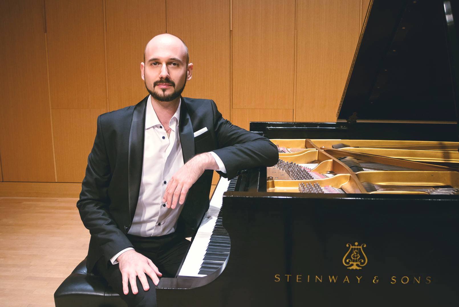 Pianist Dmitri Kristalinsky in a formal attire sitting by the concert piano facing the audience in a concert hall